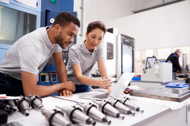 Two engineers in a factory setting examine information on a laptop. Various machine parts are spread out on the table in front of them.