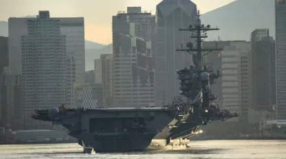 A large naval aircraft carrier sails past a city skyline with modern skyscrapers and mountains in the background.