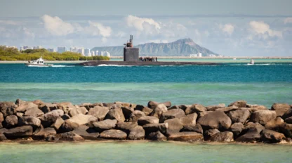 A submarine partially emerges on the ocean surface near a rocky shoreline, with a patrol boat nearby and a mountain visible in the background under a partly cloudy sky.