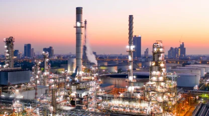 A sprawling industrial refinery with tall chimneys and complex piping, set against a city skyline at sunset.