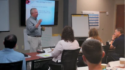 Man presenting to a group in a conference room, with a presentation slide titled "Key Learnings from Day 2" on a screen. Attendees are seated at tables, listening and taking notes.