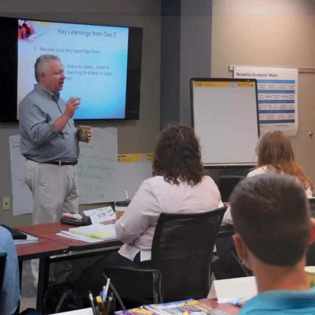 Man presenting to a group in a conference room, with a presentation slide titled "Key Learnings from Day 2" on a screen. Attendees are seated at tables, listening and taking notes.