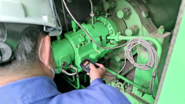 Person wearing a helmet and blue shirt uses a handheld device to inspect green industrial machinery with wires and bolts.