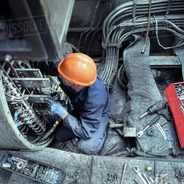 Technician in orange helmet works on electrical equipment, surrounded by tools and wires in an industrial setting.