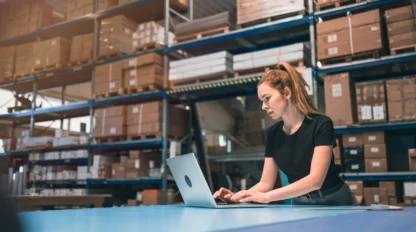 A woman in a black shirt works on a laptop in a warehouse surrounded by shelves of boxes.