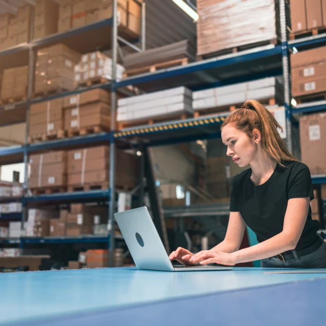 A woman in a black shirt works on a laptop in a warehouse surrounded by shelves of boxes.