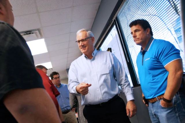 A group of people stand in a room with one man gesturing while others listen. They are near a window with blinds.