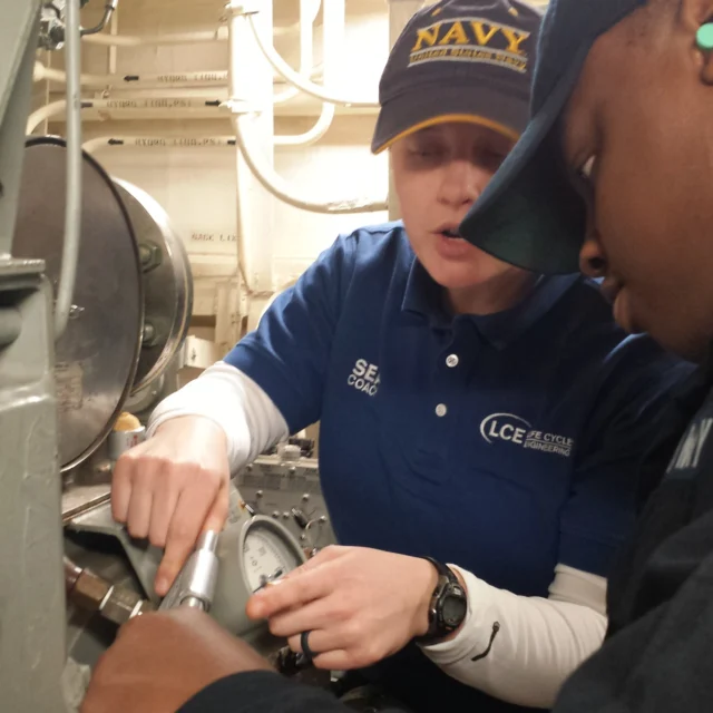 Two people in navy uniforms work together on machinery in an engine room, focusing on a gauge. One person points while the other observes closely.