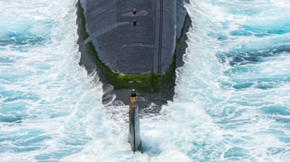 A submarine partially emerges from the ocean, surrounded by turbulent white waves and patches of green algae on its surface.