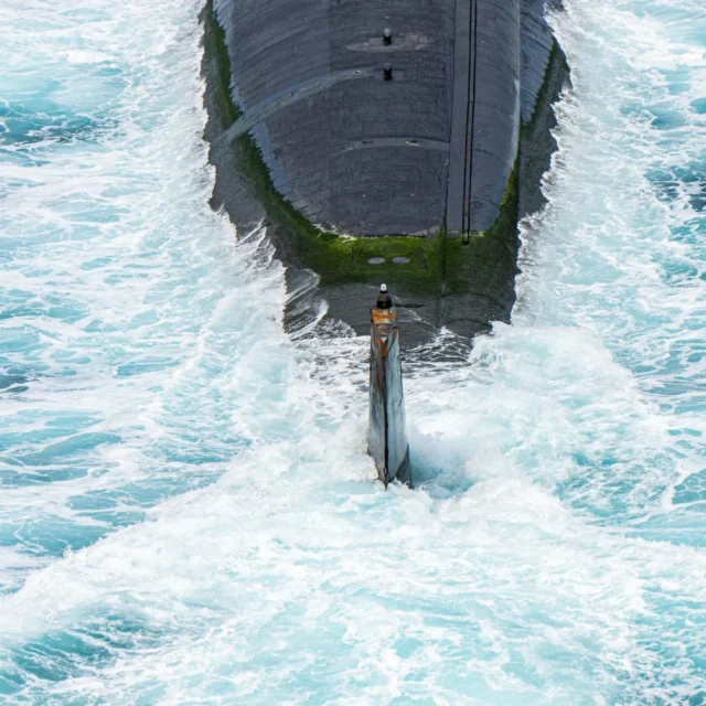 A submarine partially emerges from the ocean, surrounded by turbulent white waves and patches of green algae on its surface.