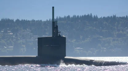 A submarine partially submerged in water, with a forested shoreline and mountains in the background under a cloudy sky.