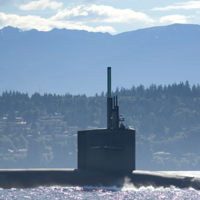 A submarine partially submerged in water, with a forested shoreline and mountains in the background under a cloudy sky.