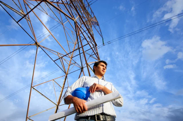 A person holding a blue hard hat and rolled-up papers stands near a tall metal power line tower against a blue sky.