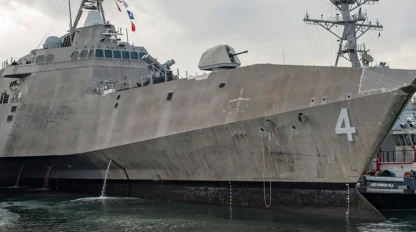 A large naval ship, marked with the number 4, docked at a harbor with other military vessels nearby. A tugboat is positioned beside it.