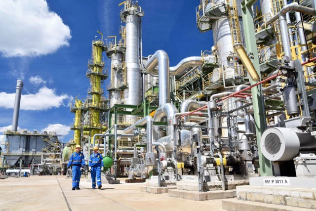 Two workers in blue uniforms and helmets walk past large industrial machinery and piping at a petrochemical plant under a blue sky.