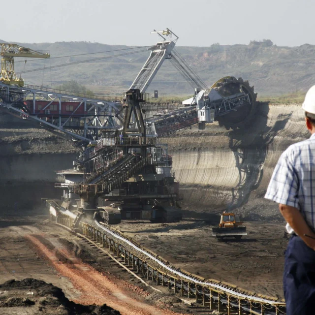 A person in a hard hat observes a large excavating machine in an open-pit mine with conveyor belts and exposed earth layers.