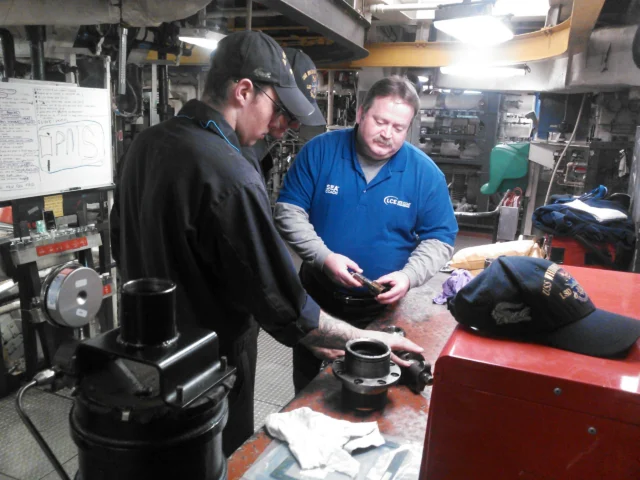 Two men work in an industrial workshop with various tools and equipment. One man inspects an object on a table. A whiteboard with notes is visible in the background.